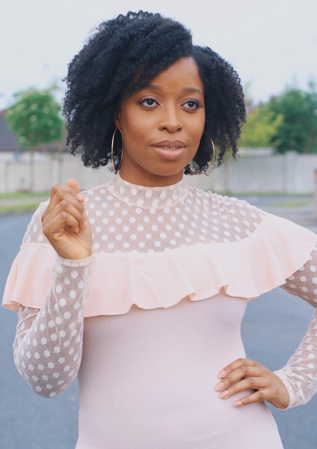 Young women wearing a pale pink dress with frills standing with one hand on her hip and the other hand raised. 
